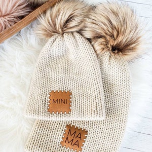 May include: Two knitted beige beanies with faux fur pom poms. The beanie on top has a brown leather patch that says "MINI" and the beanie on the bottom has a brown leather patch that says "MAMA".