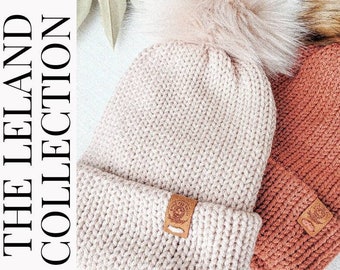 May include: Two knitted beanies with faux fur pom poms. One beanie is a light pink color and the other is a burnt orange color. Both beanies have a leather patch with a logo on the side.