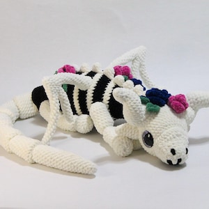 May include: A white crochet dragon with a black and white striped body, adorned with pink, blue, and green floral accents. The dragon has a long tail and is lying down. The eyes are black and the mouth is smiling.