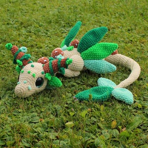 Könnte beinhalten: Ein handgehäkelter Drache in Creme-, Grün- und Brauntönen, der auf grünem Gras liegt. Der Drache hat große, runde Augen, blattförmige Flügel und einen langen, gebogenen Schwanz. Der Körper des Drachen ist mit dekorativen Elementen verziert.