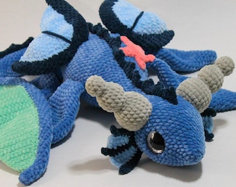 Puede incluir: Un peluche de dragón azul tejido a mano con alas y detalles en azul claro. El dragón presenta cuernos grises, un detalle en forma de estrella de mar rosa y grandes ojos negros. El peluche es suave y detallado, ideal para los amantes de la fantasía.