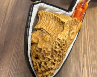 May include: A tobacco pipe carved from light tan material into the shape of a bearded face with a textured, honeycomb-like beard. The pipe features an orange, translucent stem and is housed in a black, velvet-lined case with a yellow measuring tape.