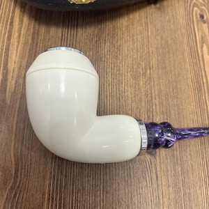 May include: Ivory-colored tobacco pipe with a curved design. The pipe features a silver band and a stem with a purple and white marbled pattern. The pipe is resting on a wooden surface, with a black case in the background.