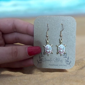 May include: A pair of gold turtle earrings with a floral design. The earrings are on a brown card with the text "Arismatic Stitches Handmade with love".