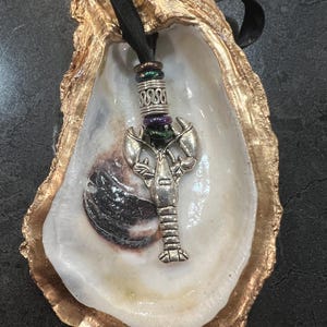 May include: A silver lobster pendant with a beaded accent hangs inside a gold-edged oyster shell. The shell is white and cream-colored, with a black ribbon for hanging. The lobster charm is detailed and the shell has a natural texture.