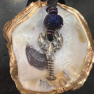 May include: An oyster shell with a gold-painted edge, holding a silver lobster charm and a dark blue beaded ornament. A black ribbon is attached for hanging. The shell's interior is white with a gray and black marking.
