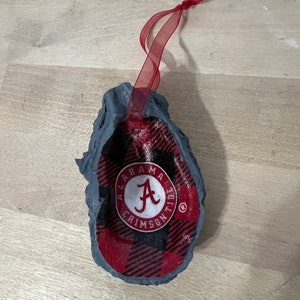 May include: A gray, textured, oyster-shaped ornament with a red and black plaid design. The ornament features a white circle with the text "Alabama Crimson Tide" and a red "A" in the center.
