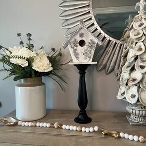May include: A decorative display featuring a white birdhouse with floral accents on a black stand, a beaded garland, a vase of white flowers, and a mirror with a silver feather design. The items are arranged on a wooden surface.