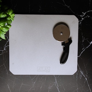 Peut inclure: Une pierre à pizza rectangulaire blanche avec un coupe-pizza noir sur le dessus. La pierre porte l'inscription "ATLAS". La pierre et le coupe-pizza sont sur une surface en marbre noir.
