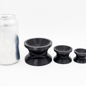 May include: Three black, 3D printed, round, bowl-shaped objects with a raised center and a criss-cross pattern on the bottom. The objects are arranged in order of size, with the largest object on the left and the smallest object on the right. A silver can is in the background.