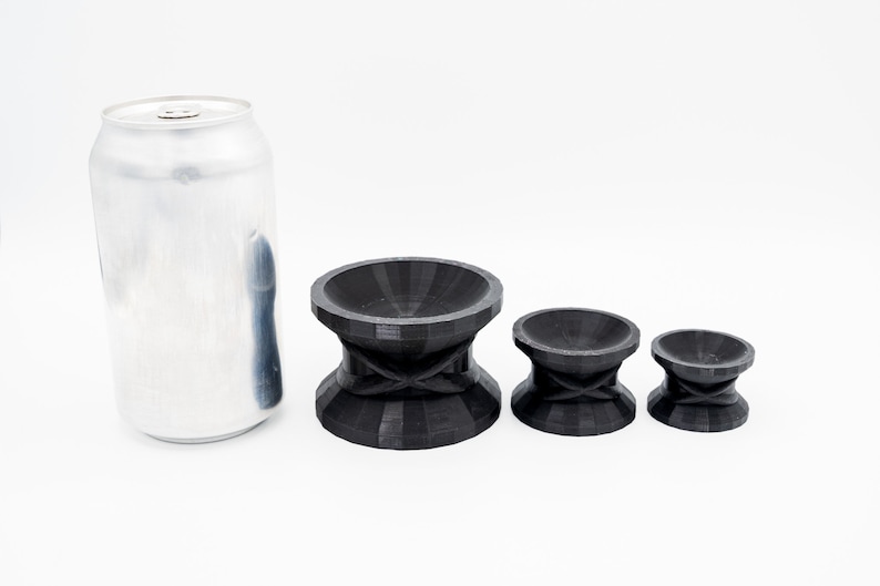 May include: Three black, 3D printed, round, bowl-shaped objects with a raised center and a criss-cross pattern on the bottom. The objects are arranged in order of size, with the largest object on the left and the smallest object on the right. A silver can is in the background.