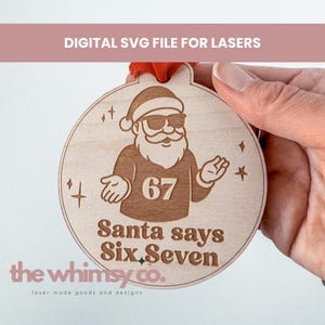 May include: Wooden Christmas ornament with a Santa Claus design wearing sunglasses and a sweater with the number 67. The text "Santa says Six, Seven" is below the Santa illustration. The ornament has a red ribbon for hanging.