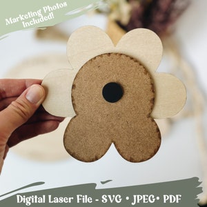 May include: A wooden flower cutout with a brown center and beige petals. The flower is held in a hand.