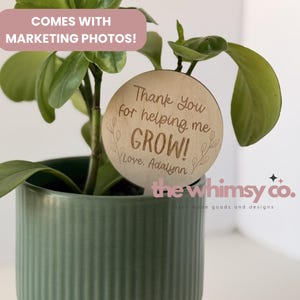 Op de afbeelding: Een groene potplant met een houten plantensteker. De steker is rond en heeft de tekst "Thank You for helping me GROW! Love, Adalynn" gegraveerd. De pot is lichtgroen en heeft verticale lijnen.