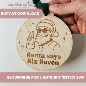 May include: A round wooden Christmas ornament featuring an engraved image of Santa Claus wearing sunglasses and giving a peace sign. The text "Santa says Six Seven" is below the image. The ornament is held by a hand.