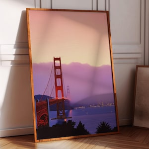 Könnte beinhalten: Ein gerahmter Druck der Golden Gate Bridge in San Francisco, Kalifornien. Die Brücke ist rot und vor einem lila und rosa Himmel platziert. Der Druck ist in einem Holzrahmen.
