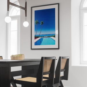 May include: A black dining table with two black chairs with woven cane seats. The table is set with a wooden bowl filled with green plants. A framed print of a blue sky, palm trees, and a swimming pool hangs on the wall behind the table.