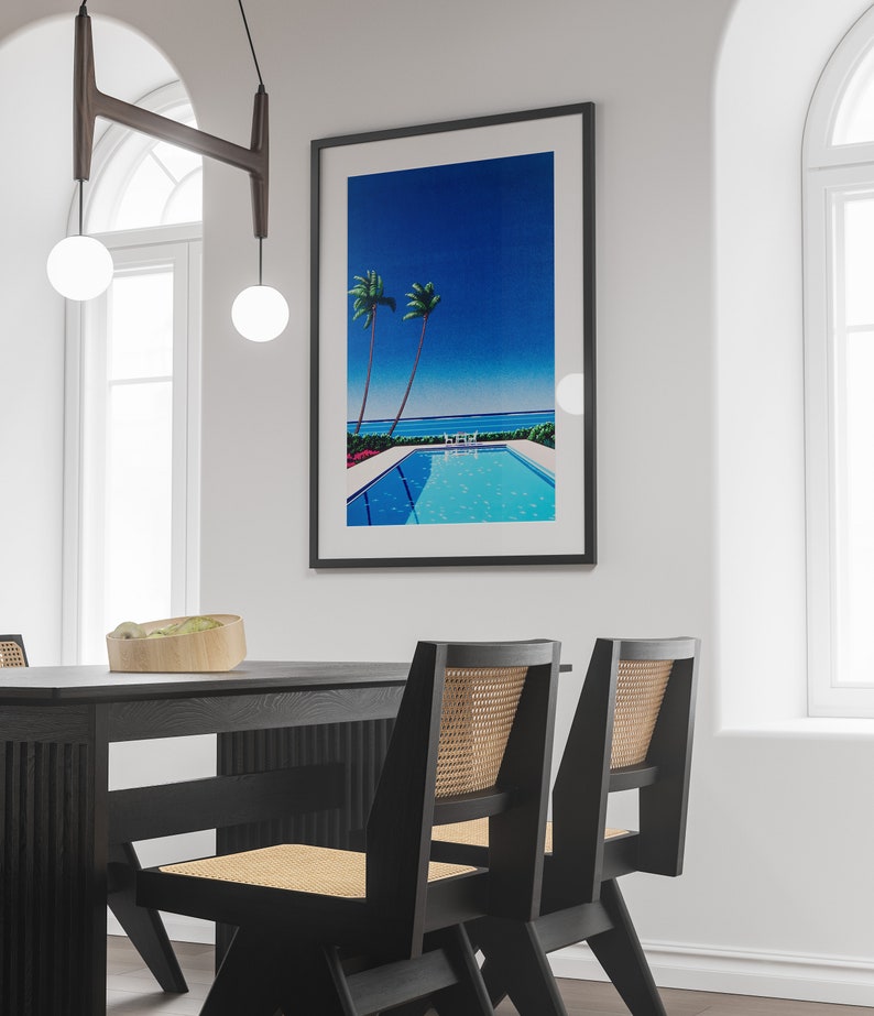 May include: A black dining table with two black chairs with woven cane seats. The table is set with a wooden bowl filled with green plants. A framed print of a blue sky, palm trees, and a swimming pool hangs on the wall behind the table.