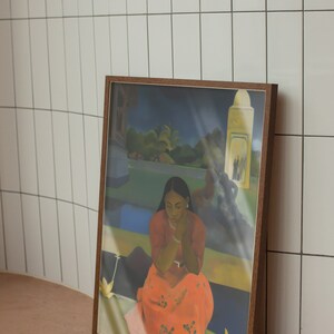 Peut inclure: Une peinture encadr&eacute;e repr&eacute;sentant une femme en robe rouge assise dans un paysage tropical. La femme regarde vers le bas et ses mains sont jointes sur ses genoux. La peinture est plac&eacute;e contre un mur en carrelage blanc.