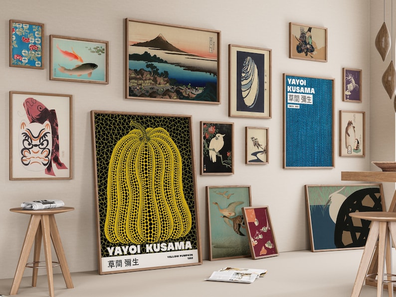 May include: A gallery wall with various framed prints, including a yellow pumpkin with black polka dots, a blue print with Japanese text, and a black and white print of a bird. The prints are arranged in a grid pattern on a white wall.
