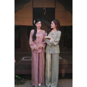 May include: Two women in matching silk pajama sets. One set is a soft pink, the other a muted green. Both have long sleeves, wide-leg trousers, and ornate gold button details. The outfits are traditional in style, reminiscent of Asian clothing.