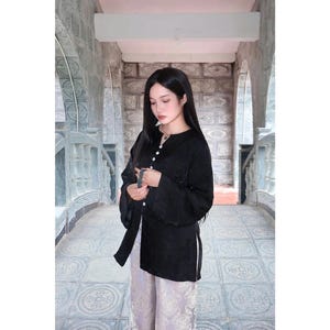 May include: A long-sleeved black tunic featuring a mandarin collar, a vertical row of small white buttons down the front, and side slits, paired with light-colored patterned trousers. The tunic has a subtle tonal texture throughout the fabric.