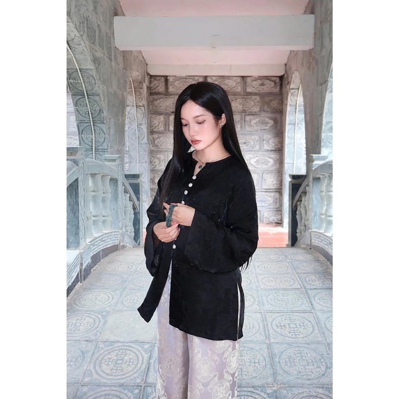 May include: A long-sleeved black tunic featuring a mandarin collar, a vertical row of small white buttons down the front, and side slits, paired with light-colored patterned trousers. The tunic has a subtle tonal texture throughout the fabric.