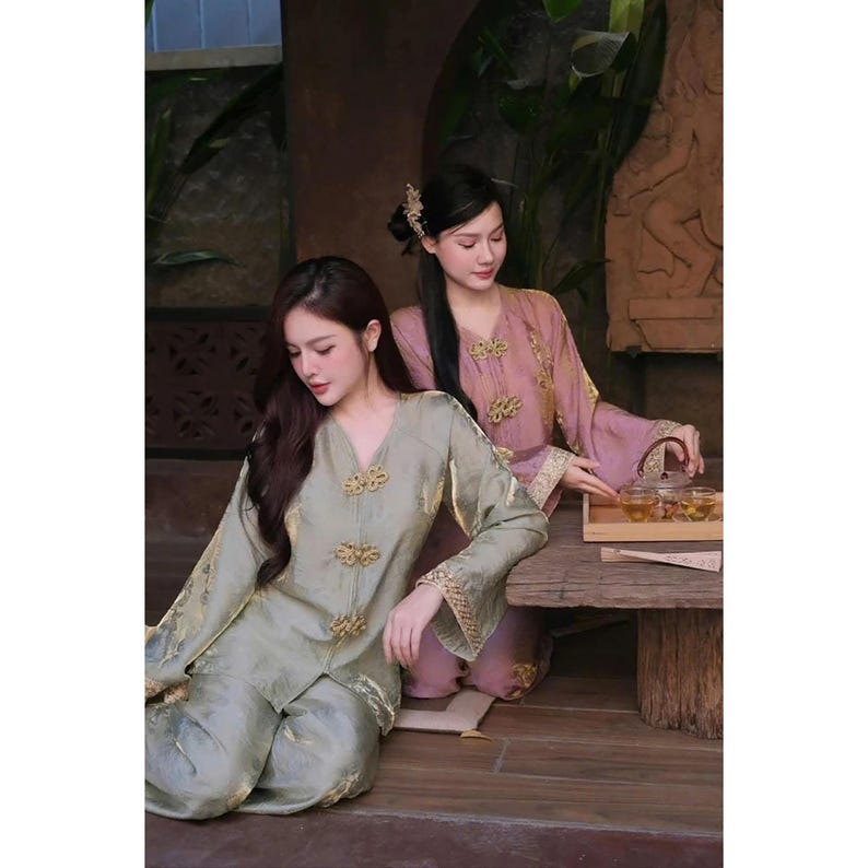 May include: Two women in traditional Asian silk outfits. One wears sage green with gold accents, the other pink. They sit near a wooden table with a tea set, suggesting a tea ceremony. The outfits have ornate button details.