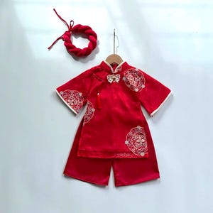 May include: A red Chinese-style outfit for a child, including a top and shorts, with white trim and embroidered details. A matching red braided bracelet is also included. The outfit is hanging on a wooden hanger.