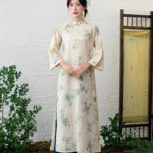 May include: A cream-coloured, long-sleeved dress with a floral pattern. The dress has a mandarin collar and a side slit. The model wears dark teal trousers and silver shoes. The background includes greenery and a wooden screen.