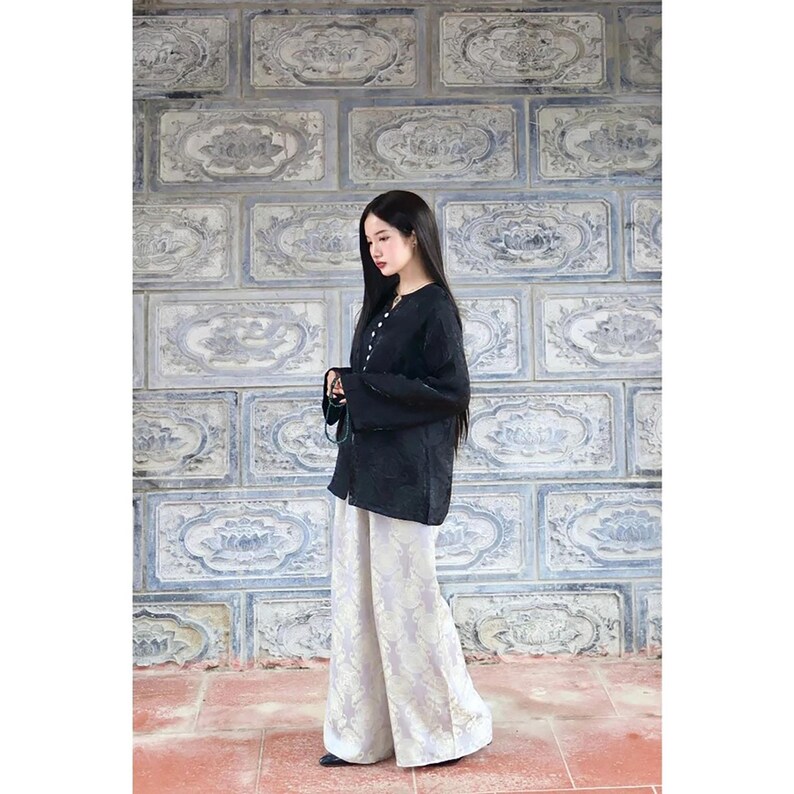 May include: A long-sleeved black top with a button-down front and a high neckline, paired with wide-leg trousers featuring a light-colored, intricate floral pattern. The ensemble consists of a dark solid-colored shirt and loose-fitting patterned pants.