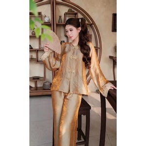 May include: Gold silk pajama set. The top features a mandarin collar and decorative frog closures. The long sleeves have embroidered cuffs. The matching trousers are wide-legged. The outfit is worn with a fan.