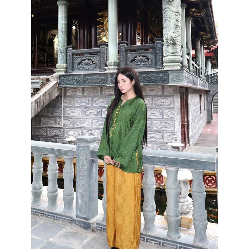 May include: A long-sleeved green tunic featuring a subtle tonal pattern and a row of gold-colored buttons along the front, paired with loose-fitting mustard yellow trousers that also display a faint, repeating tonal pattern throughout the fabric.
