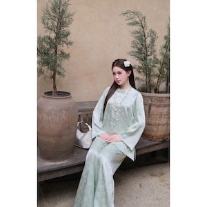 Puede incluir: Un conjunto de dos piezas de seda verde claro con estampado floral. El conjunto incluye una parte superior de manga larga y pantalones de pernera ancha. Un collar largo de perlas y un accesorio para el cabello de flores blancas completan el look. La persona lleva el conjunto en un banco de madera.
