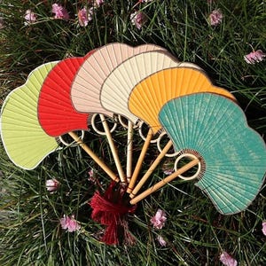May include: A collection of colorful hand fans in shades of green, red, pink, cream, yellow, and teal. The fans have bamboo handles and are adorned with a red tassel. They are arranged on a bed of green grass with pink flowers.