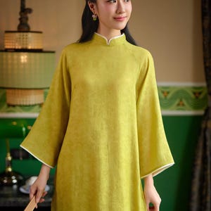 May include: A woman wearing a yellow, long-sleeved dress with a mandarin collar and white trim. The dress has a subtle pattern and is made of a soft, flowing fabric. She is holding a wooden fan in her hand.