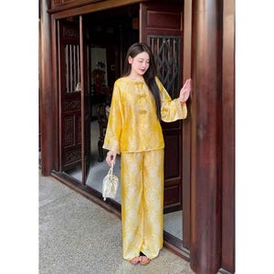 May include: A yellow silk two-piece outfit with a floral design. The long-sleeved top features decorative buttons, and the matching trousers are wide-legged. The outfit is worn with sandals and a small, white drawstring bag.