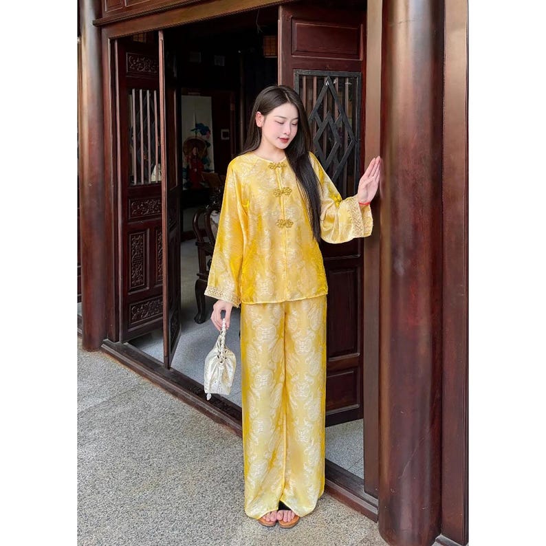 May include: A yellow silk two-piece outfit with a floral design. The long-sleeved top features decorative buttons, and the matching trousers are wide-legged. The outfit is worn with sandals and a small, white drawstring bag.