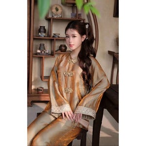 May include: A gold silk outfit, comprising a long-sleeved top with decorative button closures and matching trousers. The top has decorative trim on the cuffs. The outfit is worn by a person seated on a wooden chair.