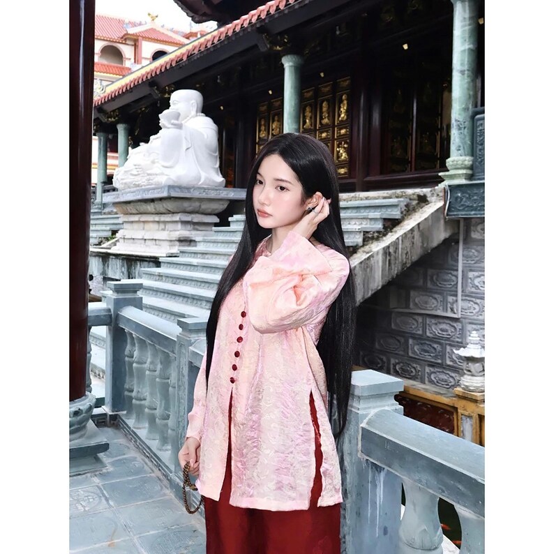 May include: A long-sleeved, light pink tunic featuring a textured, shimmering fabric with a row of small, dark red circular buttons down the front and side slits at the hem, paired with solid dark red trousers.