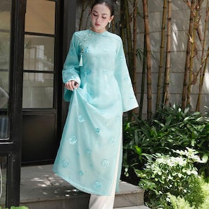 May include: A light blue, long-sleeved dress with floral embroidery and a high neckline. The dress is paired with white pants. The outfit is made of a sheer fabric and features a tassel detail near the neckline. The dress is a traditional style.
