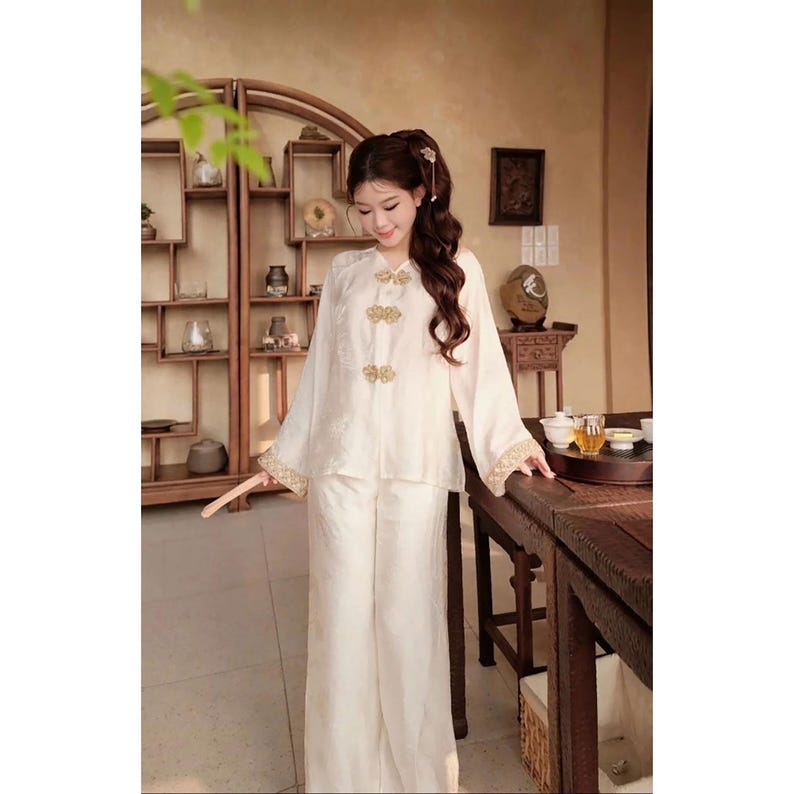 May include: A cream-coloured two-piece outfit. The long-sleeved top has gold-coloured frog closures and gold trim on the sleeves. The outfit includes wide-leg trousers. The person is in a room with wooden furniture.