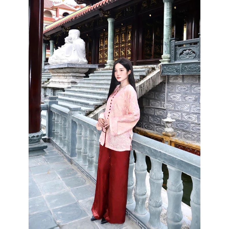 May include: A long-sleeved, light pink button-down shirt with a subtle tonal pattern paired with loose-fitting, floor-length dark red trousers. The shirt features a high collar and a vertical row of dark buttons down the front center.