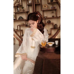 May include: A person wearing a white traditional Chinese outfit with gold trim, seated at a table with tea. The outfit features ornate gold detailing on the sleeves and front. A small tea set is on the table.