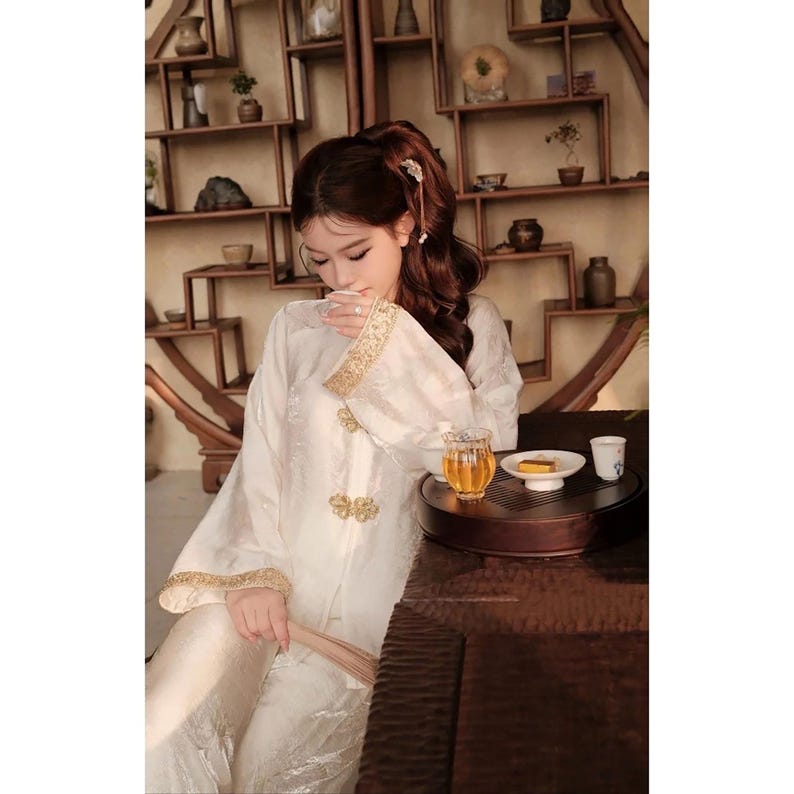 May include: A person wearing a white traditional Chinese outfit with gold trim, seated at a table with tea. The outfit features ornate gold detailing on the sleeves and front. A small tea set is on the table.