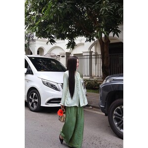 May include: A long-sleeved, light green button-down top with a textured finish paired with loose-fitting, wide-leg olive green trousers. The outfit is accessorized with a small, structured handbag featuring a bright orange flower attached to the handle.