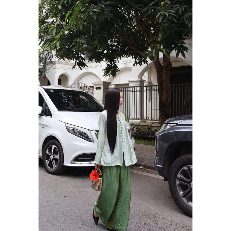 May include: A long-sleeved, light green button-down top with a textured finish paired with loose-fitting, wide-leg olive green trousers. The outfit is accessorized with a small, structured handbag featuring a bright orange flower attached to the handle.