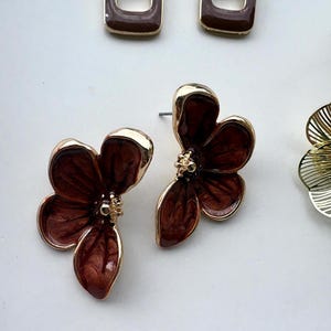 May include: A collection of earrings including two gold-toned rectangular hoops with dark brown enamel centers and two gold-toned flower-shaped stud earrings with dark brown enamel petals and a small gold center detail, plus a partial gold filigree earring.