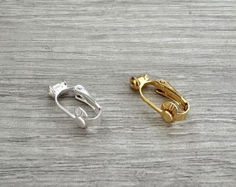 May include: Two earring findings: one silver-colored and one gold-colored. Each finding has a hinged lever-back design with a small, round post. The findings are displayed on a light gray, wood-grain surface.