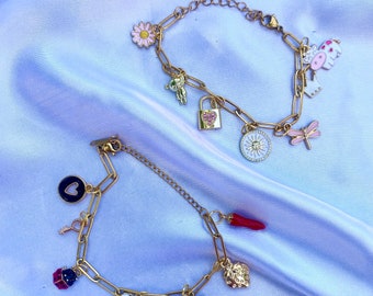 Op de afbeelding: Twee goudkleurige schakelarmbanden met bedels. De bovenste armband heeft bedels van een roze madeliefje, een teddybeer, een hartvormig hangslot, een zon, een libel en een koe. De onderste armband bevat een zwart hart, een sleutel, een lieveheersbeestje, een geblokt hart, een harten aas-kaart, een aardbei en een rode chilipeper.